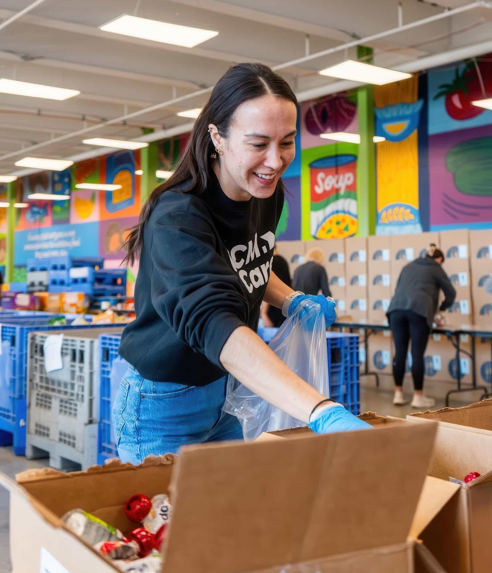 A smiling woman wearing blue gloves packs a cardboard box with food supplies at a bright, modern community food bank.